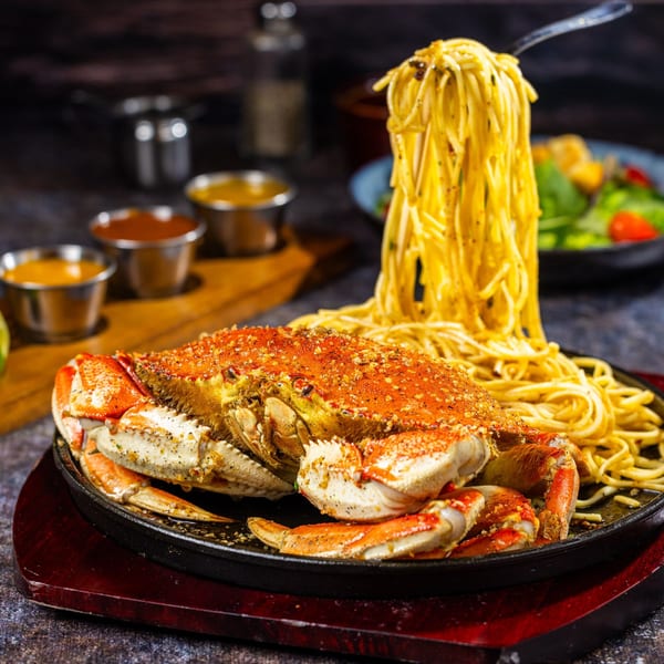 A whole roasted crab with a pile of spaghetti and a fork lifting noodles, served on a black skillet.