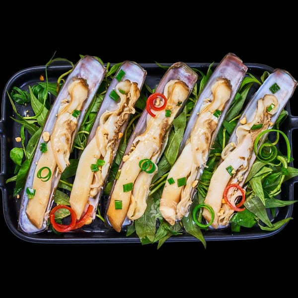 Five razor clams in their shells, garnished with herbs, peanuts, and chili, on a black platter.