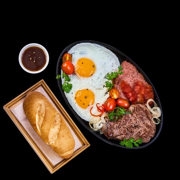 A breakfast skillet with fried eggs, steak, sausage, tomatoes, onions, and a baguette with sauce.