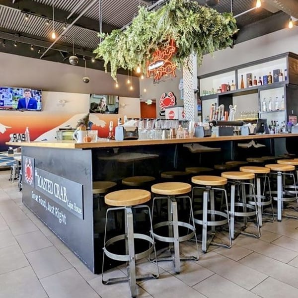 The Roasted Crab restaurant bar with stools, a neon crab sign, and hanging greenery.