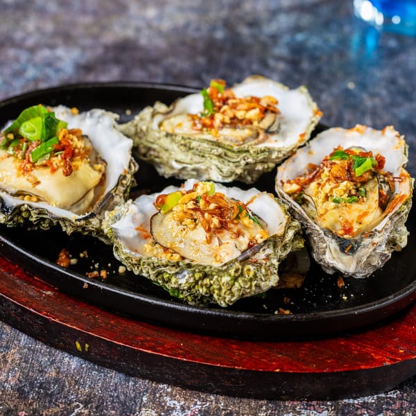 Four baked oysters topped with green onions and crispy garlic, served on a black sizzling plate.